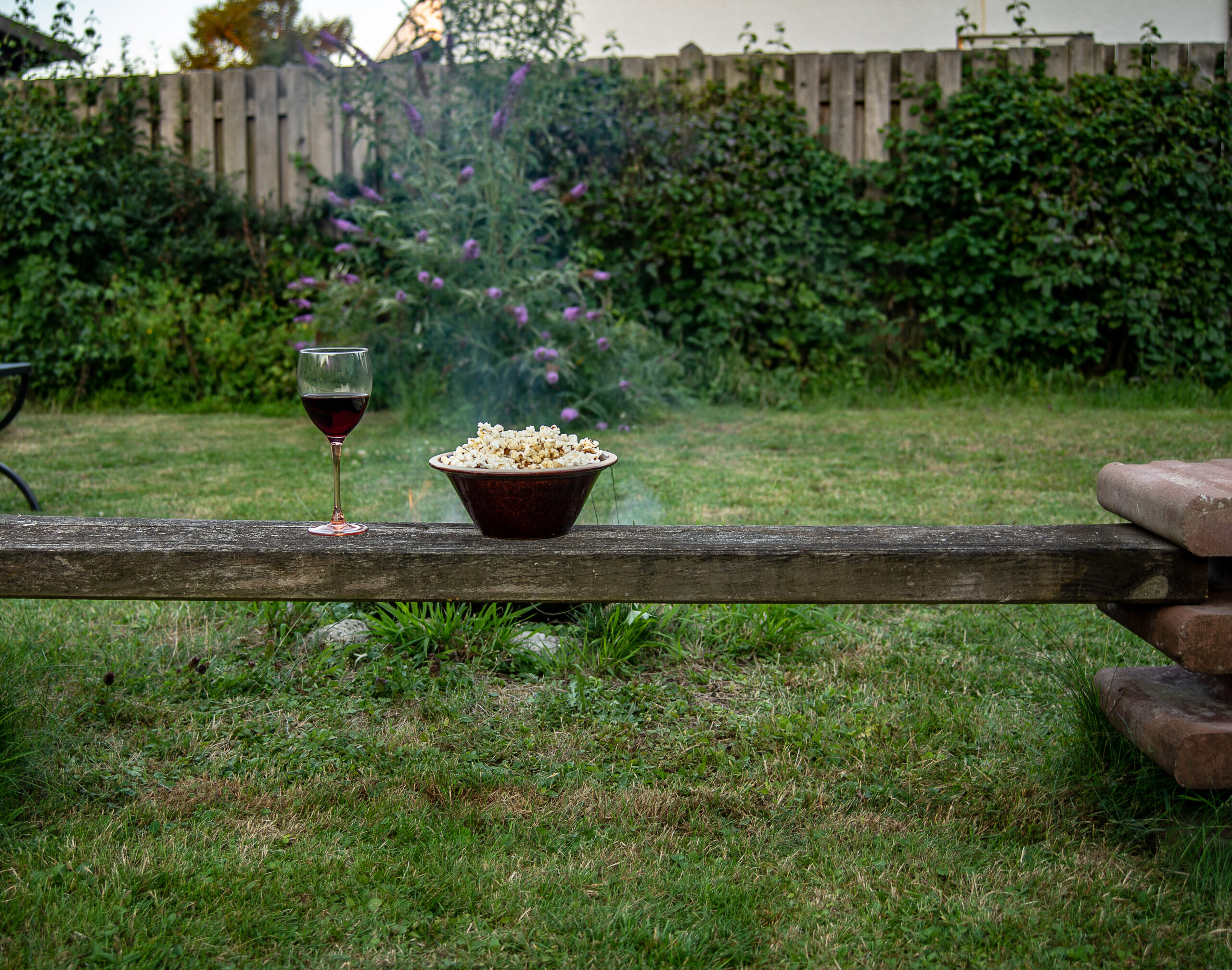 Bowl of popcorn and glass of wine stand on a bench with a campfire behind - feature image for campfire popcorn