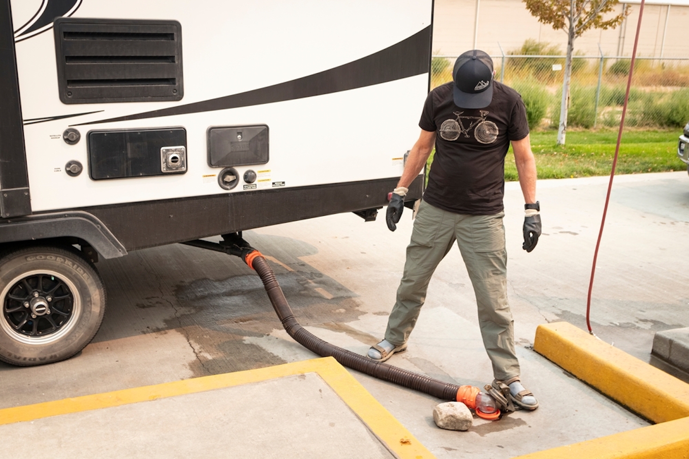 Man in tshirt and baseball cap stands between RV sewer hose and RV at sanidump - feature image for RV gray water tank cleaning