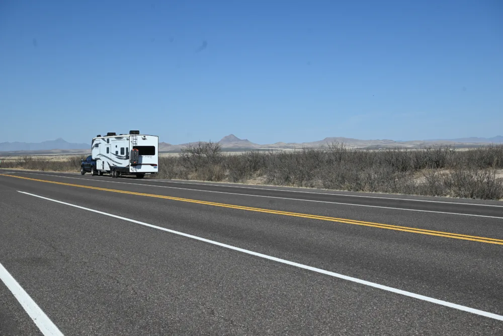 a white truck driving down a highway next to a dry grass and brush covered hill covered in a blue sky