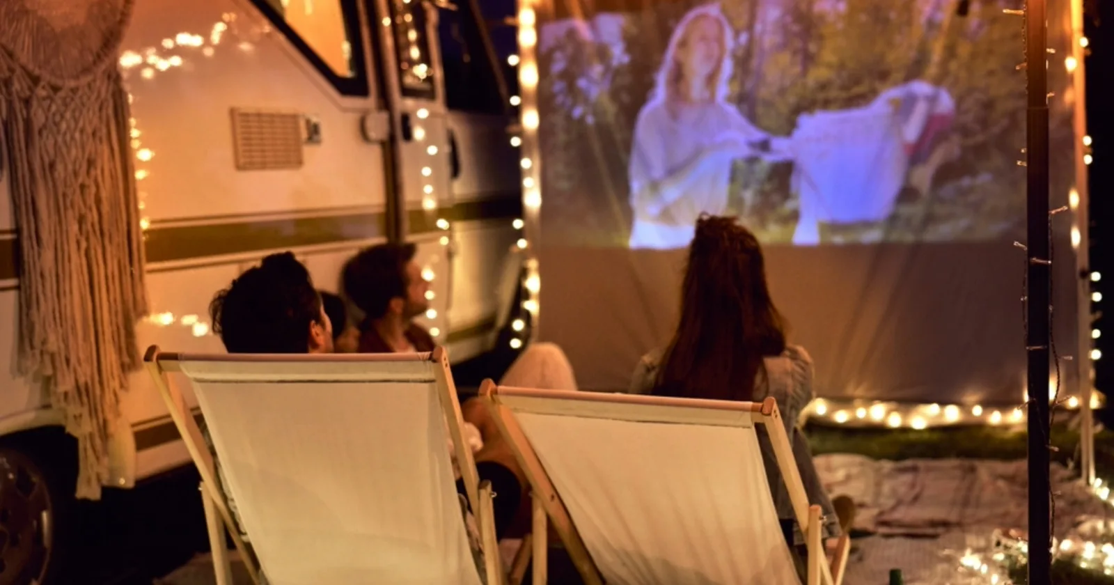 Young cheerful people watching a movie outdoors by their RV on camping site during RV movie night