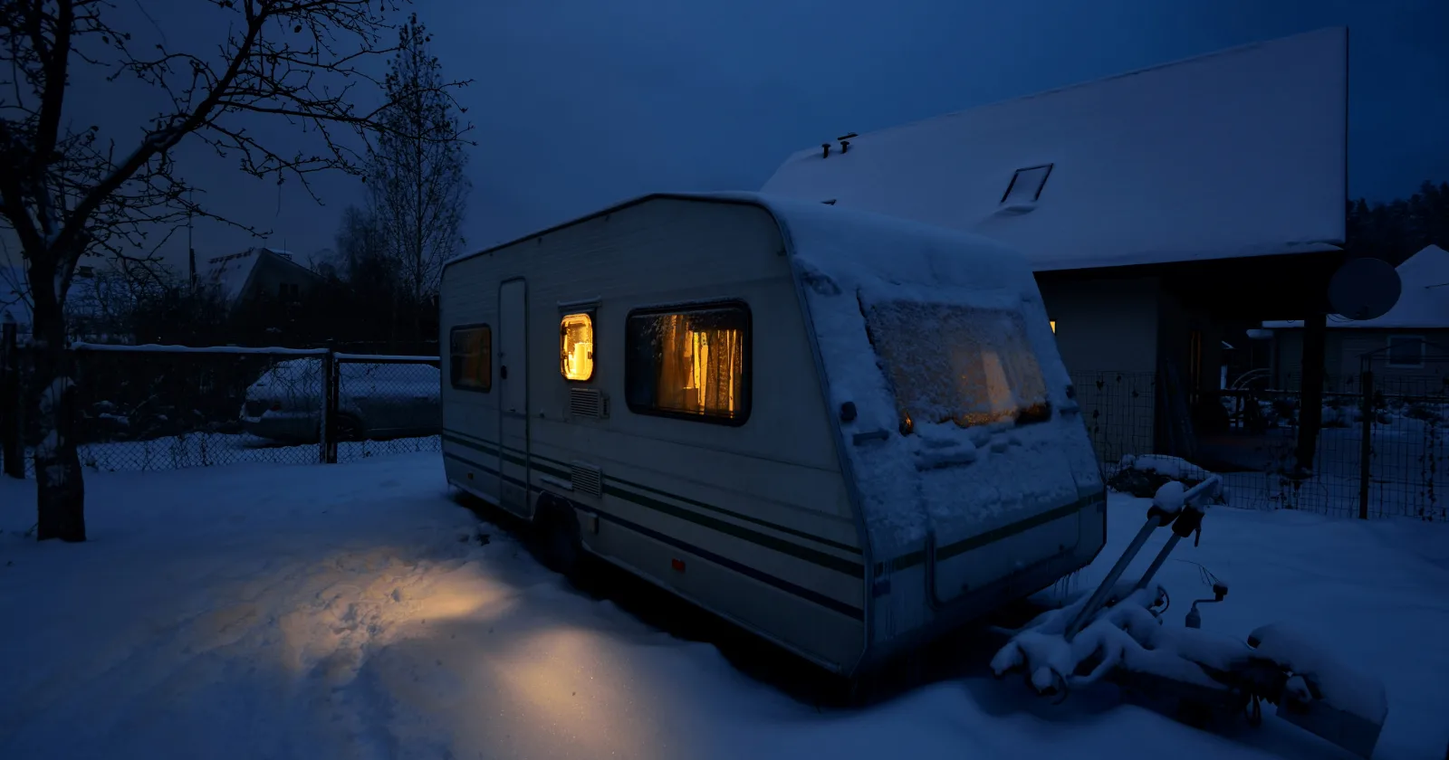 Snow covered travel trailer with lights on inside parked next to house and not being used till camping season