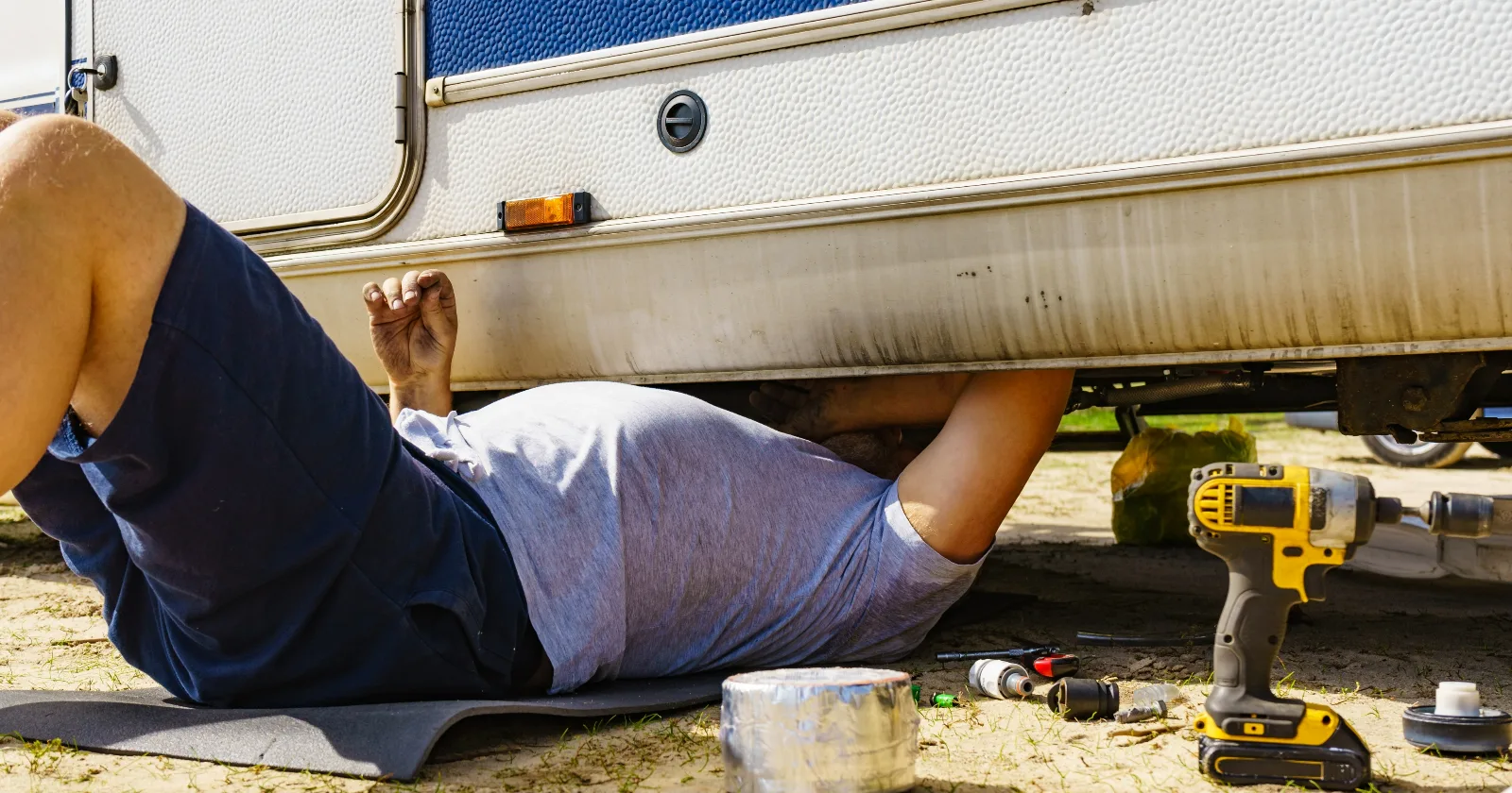 Man lying on ground, repairing bottom of the caravan vehicle with tools lying around