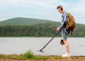 man using metal detector on shore of lake near forest as one of his RV hobbies